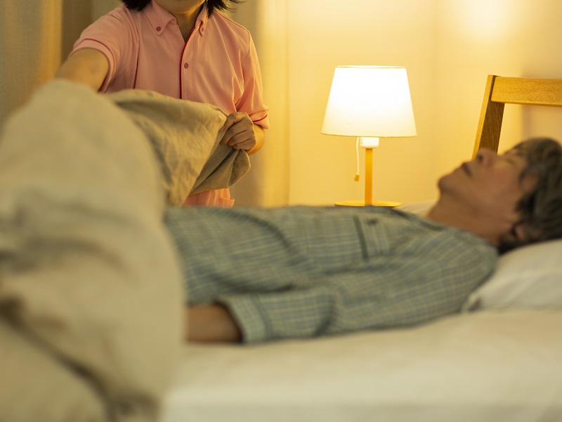 ベッドに横たわる男性に女性が布団を掛けている様子