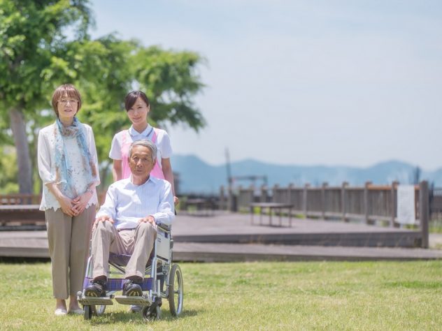 kn_390 車椅子の高齢男性と隣に立つ高齢女性と車椅子を押す女性介護スタッフの画像