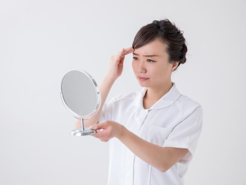 前髪を触りながら鏡を見て、考え込んでいる様子の看護助手の女性のイメージ