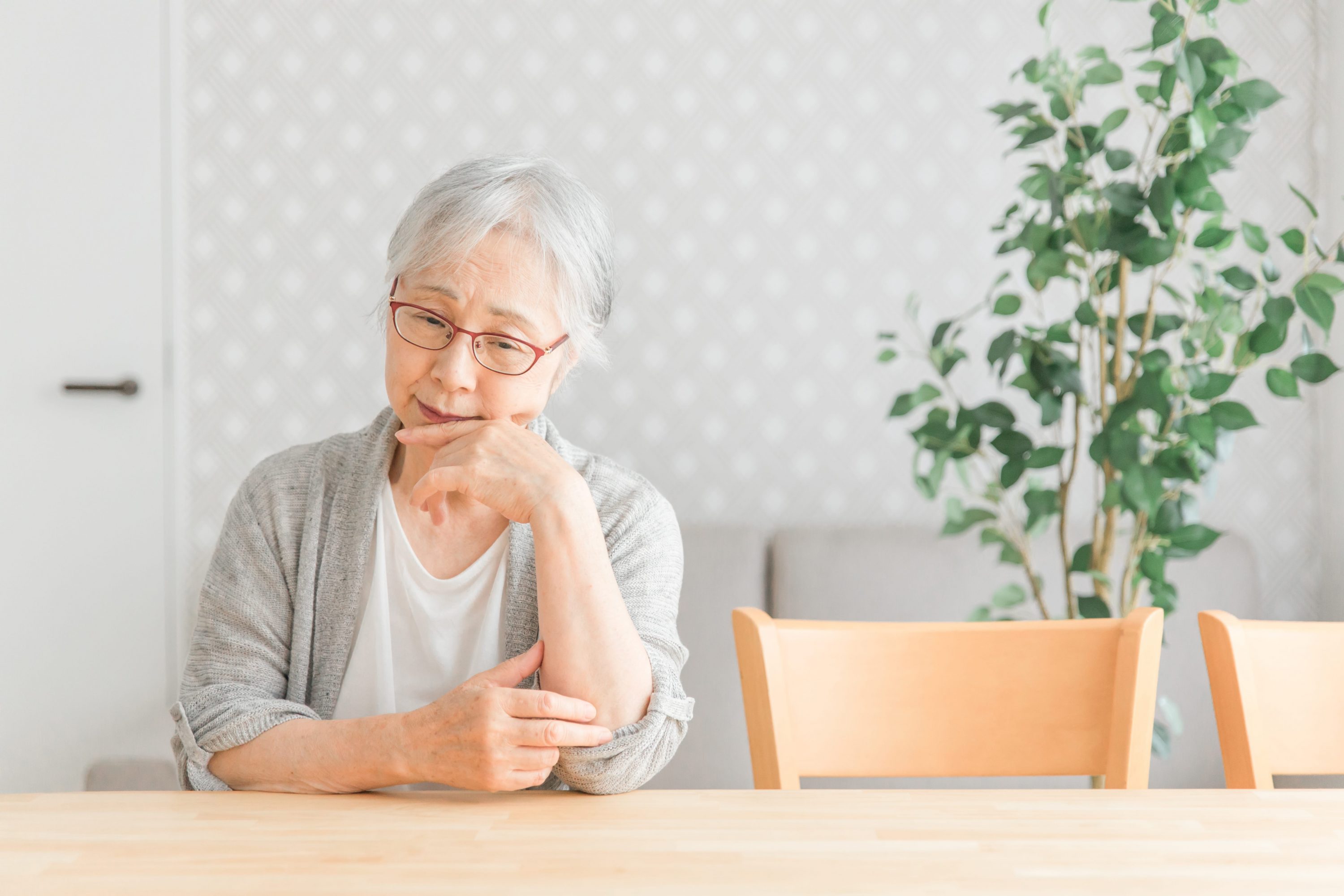 悩んだ様子で机の上で肘をついている女性高齢者のイメージ
