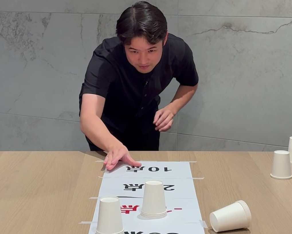 点数が書かれた紙に向かって紙コップを滑らせている男性の画像